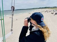 Eine Frau steht auf einem Wachturm und guckt durch ein Fernglas auf den Strand. Auf dem Strand sind Menschen und Meer in der Ferne zu erkennen.